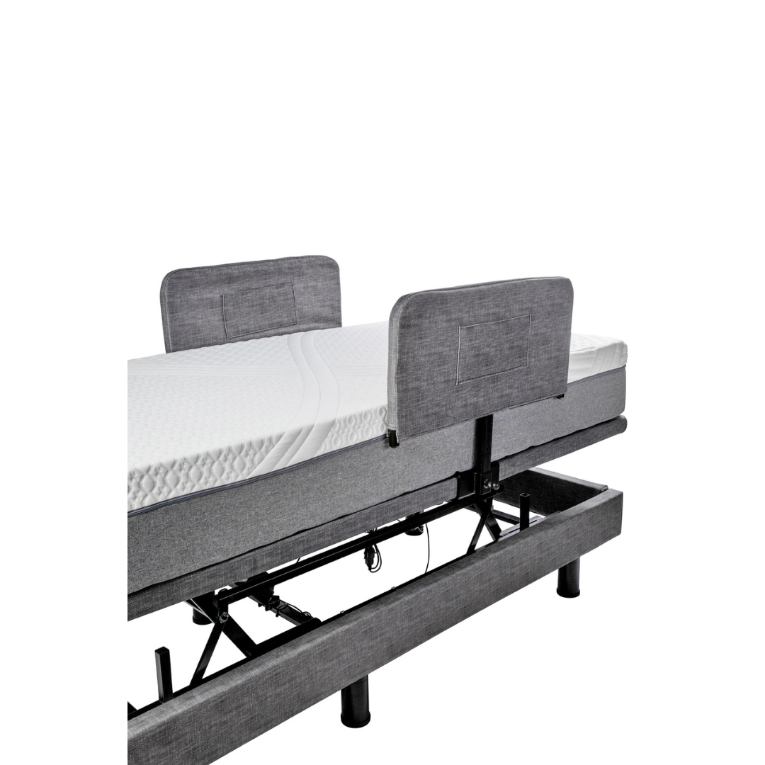 The Golden Tech Passport Homecare Bed features a gray fabric upholstered headboard, adjustable base with a white mattress, and raised frame to expose mechanical components, blending modern style with essential accessories for enhanced safety.